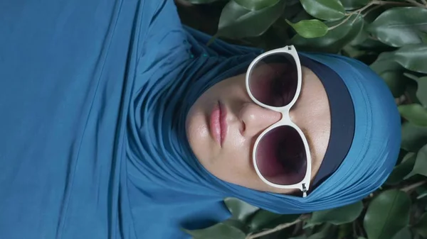 Retrato de una hermosa mujer musulmana caucásica con gafas de sol sobre un fondo de follaje ficus, vertical — Foto de Stock