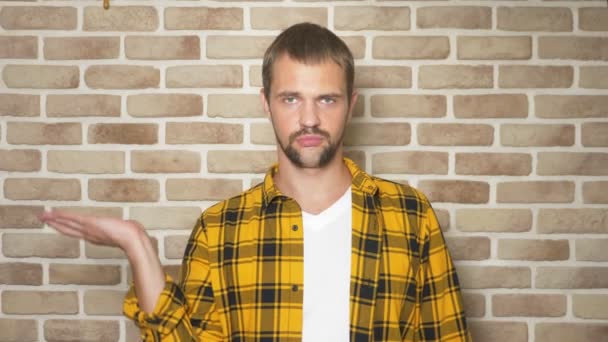 El hombre guapo en la camisa a cuadros a la moda amarilla compara, las variantes para los pesos, contra la pared de ladrillo — Vídeo de stock