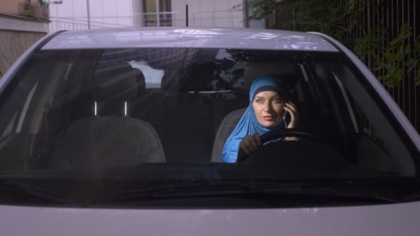Вид через лобовое стекло автомобиля. Мусульманка в синем хиджабе разговаривает по телефону за рулём машины . — стоковое видео