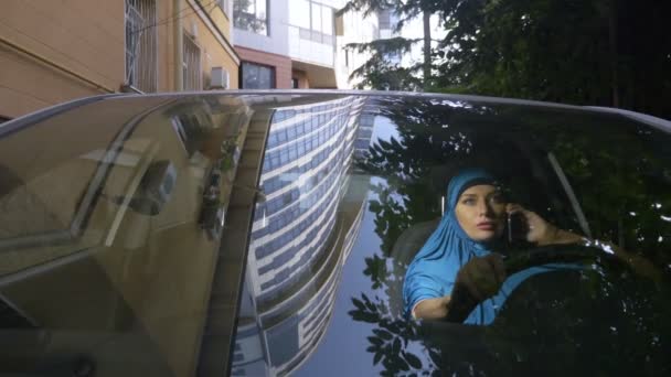 Zicht door de voorruit van een auto. Moslim vrouw in blauwe hijab praten op de telefoon tijdens het rijden van een auto. — Stockvideo