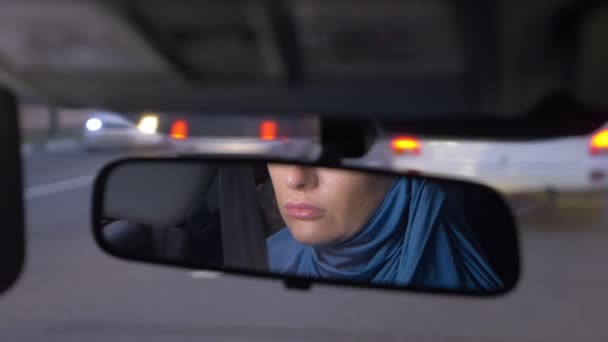 Reflejo de una mujer en un hiyab en un espejo retrovisor de un coche mientras conduce en un atasco de tráfico por la noche. Mujer musulmana conduciendo un coche . — Vídeo de stock