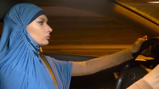 Vacker muslimsk kvinna i en blå hijab rider en bil på kvällen genom gatorna i staden. — Stockvideo