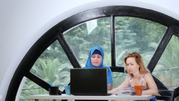 Conceito de equipa multinacional. Duas mulheres, uma muçulmana num hijab e uma caucasiana num top aberto, trabalham juntas no escritório usando um portátil. . — Vídeo de Stock
