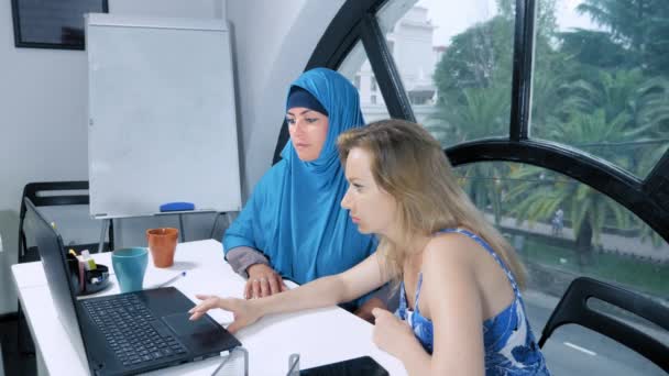 Çok uluslu takım konsepti. İki kadın, başörtüsü takan Müslüman bir kadın ve üstü açık beyaz bir kadın, ofiste dizüstü bilgisayar kullanarak birlikte çalışıyor.. — Stok video