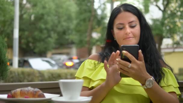 Krásná brunetka používající mobilní telefon v kavárně venku. Snídaně s croissantem a kávou, která kontroluje Novinky ve vašem smartphone. — Stock video