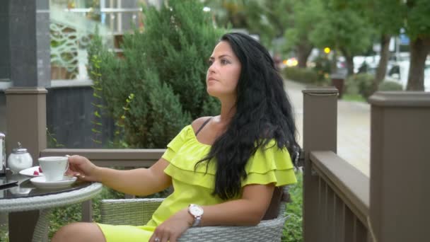 Hermosa chica morena con el pelo largo sentado mientras desayunaba en un café al aire libre, tomando café con un croissant y descansando — Vídeos de Stock