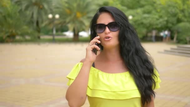 Giovane bella ragazza bruna sorride quando cammina nel parco con il suo smartphone. donna che parla al telefono all'aperto — Video Stock