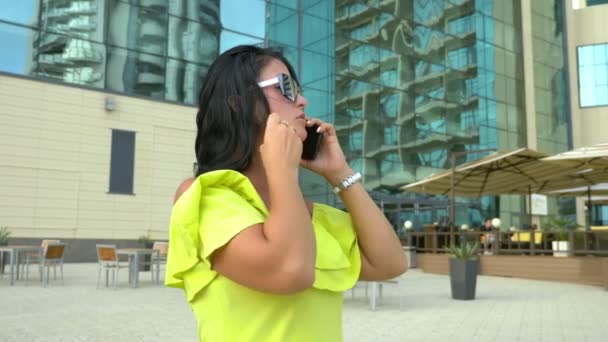 Bella giovane donna bruna che cammina attraverso il centro della città utilizza uno smartphone. giornata estiva soleggiata. stile di vita di successo. Ritratto femminile — Video Stock