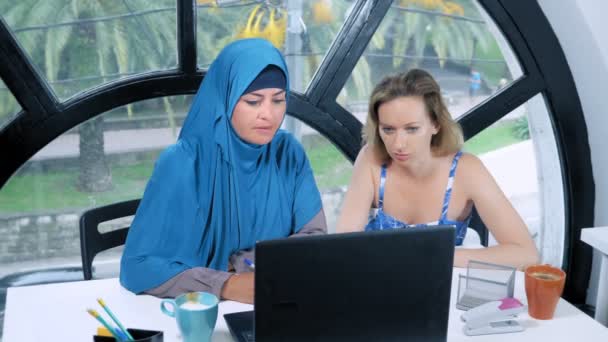 Multinationale team concept. Twee vrouwen, een moslim vrouw in een hijab en een blanke vrouw in een open top, werken samen op kantoor met behulp van een laptop. — Stockvideo