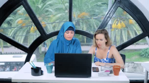 Multinationale team concept. Twee vrouwen, een moslim vrouw in een hijab en een blanke vrouw in een open top, werken samen op kantoor met behulp van een laptop. — Stockvideo