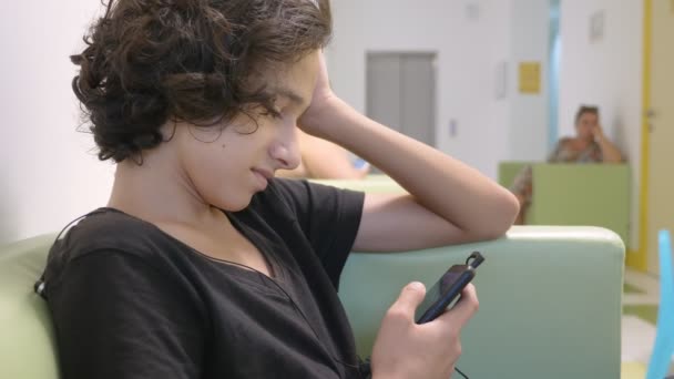 Adolescent care stă în holul unei clinici pentru copii, așteptând o întâlnire cu un medic, folosind un smartphone. Conceptul de examinare medicală, monitorizare a sănătății, examinare preventivă a — Videoclip de stoc