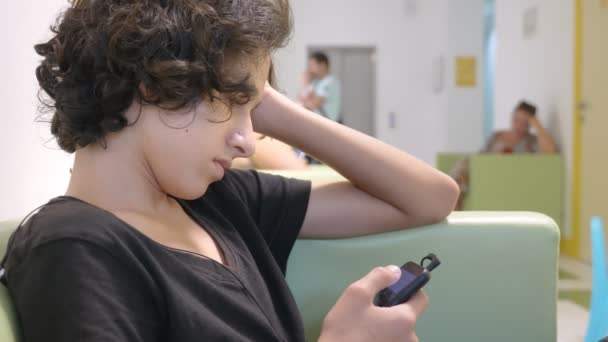 Adolescent assis dans le hall d'une clinique pour enfants, attendant une réunion avec un médecin, à l'aide d'un smartphone. Le concept d'examen médical, de suivi de la santé, d'examen préventif des — Video