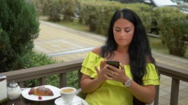 Güzel esmer kadın açık havada kahve dükkanında cep telefonu kullanarak. Kruvasan ve kahve ile kahvaltı, akıllı telefonunuzdaki haberleri kontrol edin.