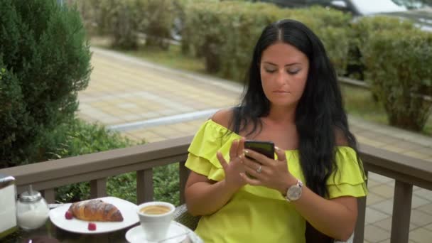 Hermosa mujer morena utilizando el teléfono celular en la cafetería al aire libre. Desayuno con un croissant y café, revisa las noticias en tu smartphone . — Vídeo de stock