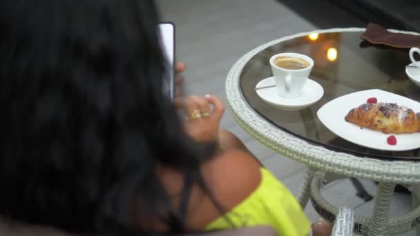 屋外のコーヒーショップで携帯電話を使用して美しいブルネットの女性。クロワッサンとコーヒーを使った朝食、スマートフォンでニュースをチェック. — ストック動画
