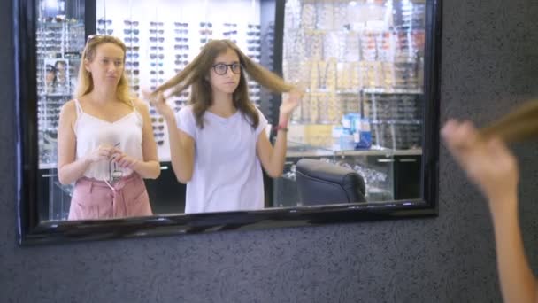 Escolha de armações de copos em óptica. Mãe e sua filha adolescente escolhem uma moldura para óculos enquanto olham em um grande espelho na óptica . — Vídeo de Stock