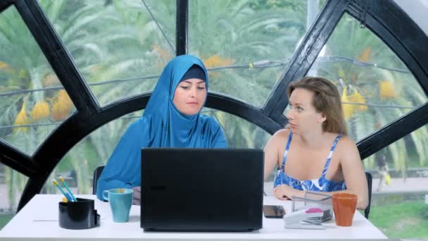 Conceptul de echipă multinațională. Două femei, o femeie musulmană într-un hijab și o femeie caucaziană într-un top deschis, lucrează împreună la birou folosind un laptop. . — Videoclip de stoc