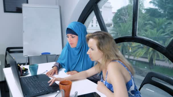 Concetto di squadra multinazionale. Due donne, una musulmana in hijab e una caucasica in open top, lavorano insieme in ufficio usando un portatile . — Video Stock