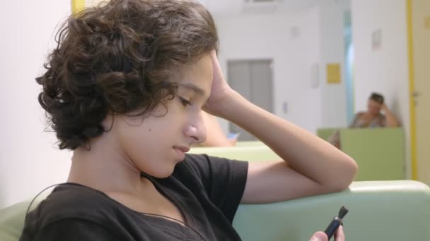 Adolescente sentado en el vestíbulo de una clínica infantil, esperando una reunión con un médico, usando un teléfono inteligente. El concepto de examen médico, vigilancia de la salud, examen preventivo de — Vídeos de Stock