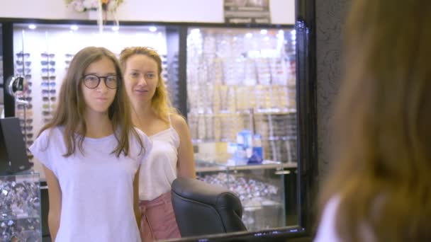 Choix de montures pour lunettes en optique. Maman et sa fille adolescente choisissent un cadre pour les lunettes tout en regardant dans un grand miroir en optique . — Video