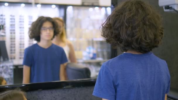 Auswahl von Fassungen für Brillen in Optik. Mutter und ihr Teenager-Sohn wählen Brillengestell, während sie in einen großen Spiegel in Optik schauen. — Stockvideo