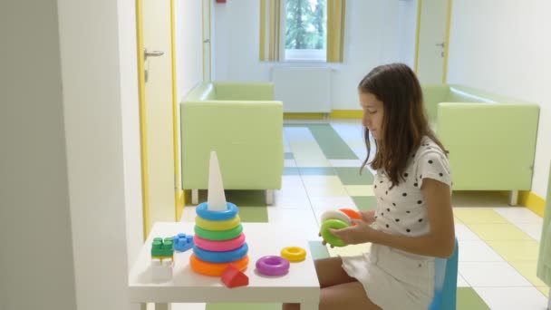 Una chica se sienta en el vestíbulo de una clínica infantil, esperando una reunión con un médico, ordenando una pirámide infantil. El concepto de examen médico, vigilancia de la salud, examen preventivo de — Vídeos de Stock