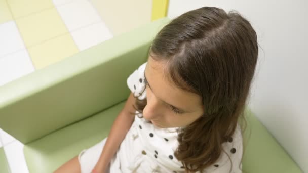 Dziewczyna siedzi w holu kliniki dziecięcej, czekając na wizytę u lekarza. koncepcja badania lekarskiego, monitorowanie stanu zdrowia, profilaktyczne badanie lekarzy. — Wideo stockowe