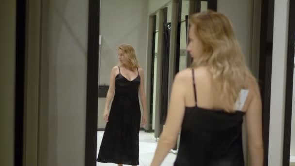 Nő, hosszú hajú próbál egy hosszú, Fekete estélyi ruha mellett a tükör egy illeszkedő helyiségben egy butik áruház — Stock videók