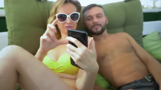 Ein Mann und eine Frau im Badeanzug rauchen eine Wasserpfeife, trinken Cocktails in einem Pavillon am Strand und benutzen ein Smartphone — Stockvideo