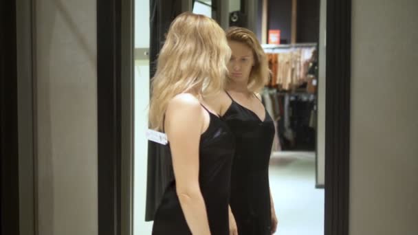 Женщина с длинными волосами примеряет длинное черное вечернее платье рядом с зеркалом в примерочной бутик-магазина — стоковое видео