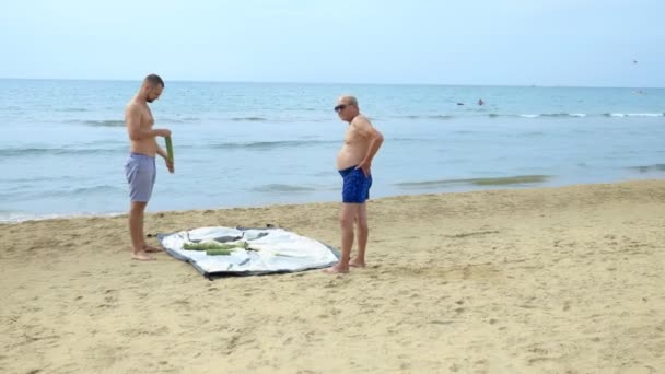 Мужчина и его отец разбили палатку для кемпинга на пляже на голубом морском фоне летом — стоковое видео