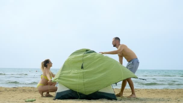 Par, man och kvinna inrätta ett tält för Camping på en sandstrand nära havet på en varm solig dag — Stockvideo