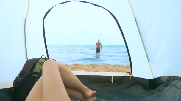 Camping concept aan zee. uitzicht vanuit de tent, het meisje kijkt naar haar vriendje spetteren in de zee, liggend in de tent — Stockvideo