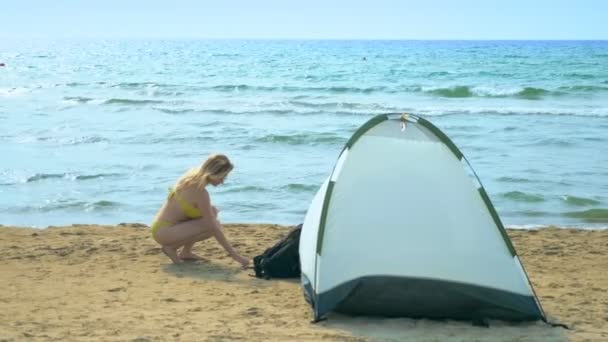 Camping konceptet vid havet. Flickan samlar snäckor på stranden nära tältet på en bakgrund av havet. — Stockvideo