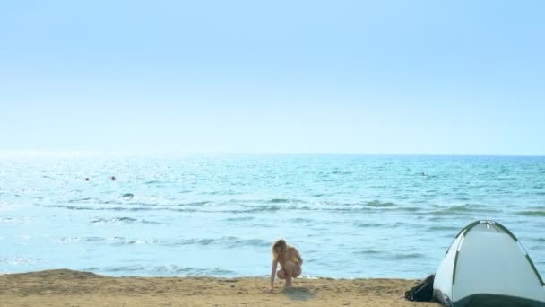 Concepto de camping junto al mar. Chica recoge conchas en la playa cerca de la tienda de campaña en un fondo del mar . — Vídeos de Stock