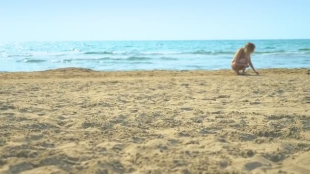 Vrouw in een badpak wandelingen langs de kust, verzamelt schelpen op een zandstrand. — Stockvideo