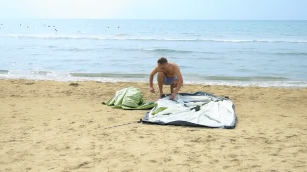 Chico en pantalones cortos establece una tienda de campaña para acampar en una playa de arena con un fondo de mar en el verano — Vídeo de stock