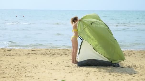 Chica en traje de baño pone una tienda de campaña para acampar en una playa de arena contra el mar en el verano — Vídeos de Stock