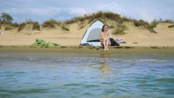Una chica en traje de baño está descansando junto a una tienda de campaña en una playa de arena contra el fondo del mar y las dunas en el verano. concepto de unidad con la naturaleza — Vídeos de Stock