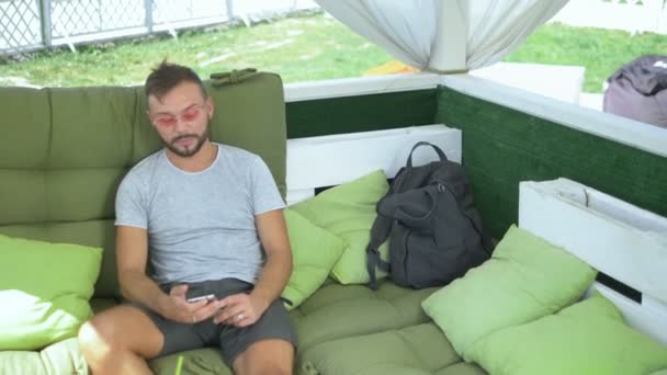 Handsome guy uses a smartphone while relaxing on a gazebo bed — Stock Video