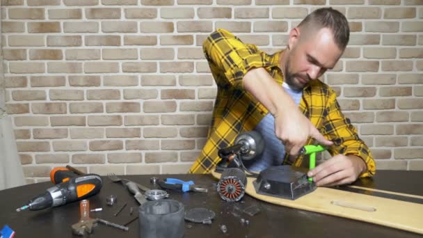 Ένας άντρας μηχανικός επισκευάζει ένα ηλεκτρικό σκέιτ που κάθεται σε ένα εργαστήρι σε μια σοφίτα. κύριο σχέδιο — Αρχείο Βίντεο