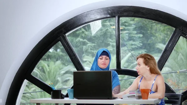 multinational team concept. Two women, a Muslim woman in a hijab and a Caucasian woman in an open top, work together at the office using a laptop.
