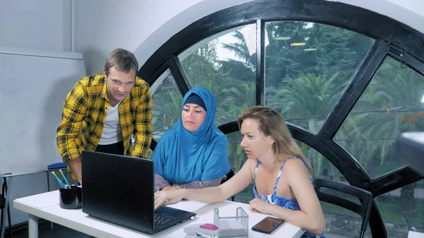 Concept d'équipe multinationale. femme musulmane en hijab, femme caucasienne et homme caucasien travaillant ensemble dans le bureau à l'aide d'un ordinateur portable . — Photo