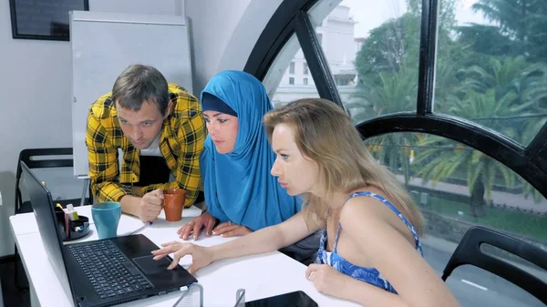 多国籍チームのコンセプト。ヒジャーブのイスラム教徒の女性、白人女性と白人男性は、ラップトップを使用してオフィスで一緒に働く. — ストック写真