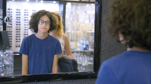 Elección de monturas para gafas en óptica. Mamá y su hijo adolescente eligen un marco para gafas mientras se miran en un espejo grande en óptica . — Foto de Stock