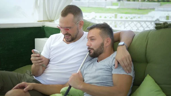 Um casal gay fuma um narguilé e observa o aplicativo no telefone, sentado no gazebo na rua em um dia ensolarado de verão . — Fotografia de Stock