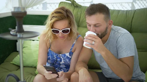 couple man and woman smoke a hookah in the gazebo on a clear sunny day and use a smartphone