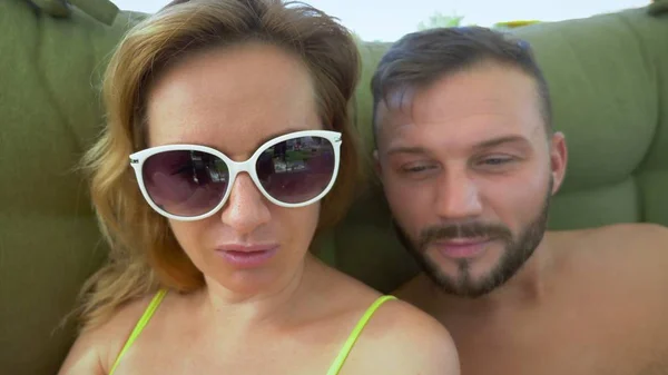 Um homem e uma mulher de fato de banho fumam um cachimbo, bebem cocktails num gazebo na praia e usam um smartphone — Fotografia de Stock