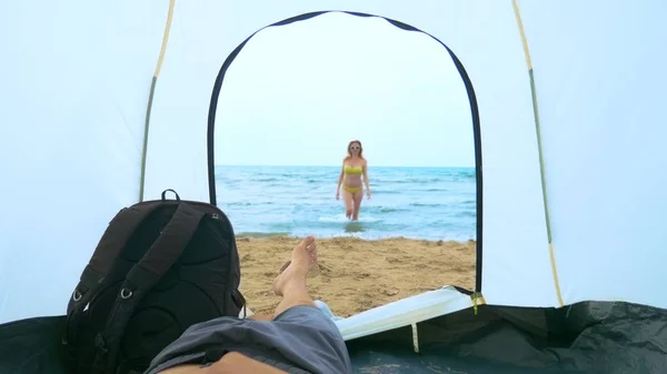 Concepto de camping junto al mar. un hombre mira desde una tienda de campaña para su novia que salpica en el mar — Foto de Stock