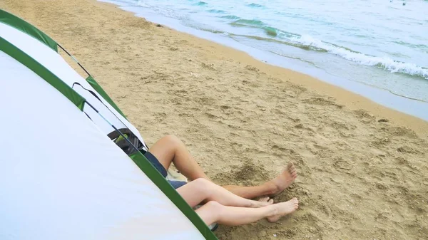 Concepto de camping junto al mar. las piernas de un hombre y una mujer son visibles desde una tienda de campaña sobre la arena sobre el fondo del mar . — Foto de Stock